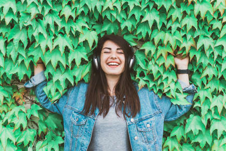 Young hipster girl listens to music leaning against an ivy wall - Pretty woman relaxes with headphones in a city parkの写真素材