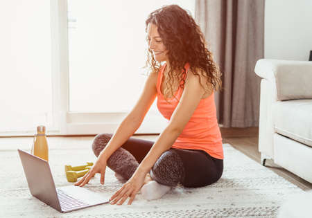 Young woman in sporty outfit does exercises with dumbbells at home watching tutorial on laptop -...