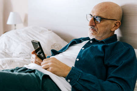 Middle-aged man rests on the bed with smart phone - Handsome man reads latest news and does online shopping from mobile devices in leisure time - elderly and technology conceptの写真素材