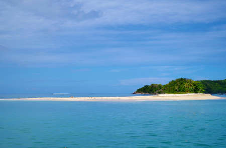 A shot of a very calm sea on a sunny day. There are few scattered clouds in the sky. On the horizon we can see a small island, overgrown with palm trees and sporting an inviting, sandy beachの写真素材