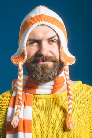 Portrait of smiling bearded man with scarf and hat on blue background. Young handsome man wearing...