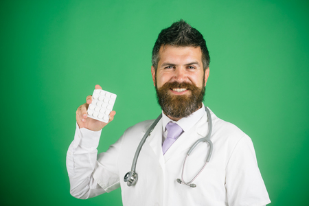 Portrait of young male doctor with of pills. Physician with beard dressed in white medical robe with pill. Medicine and health concept. Physician with stethoscope in medical gown with pill in hand.の写真素材