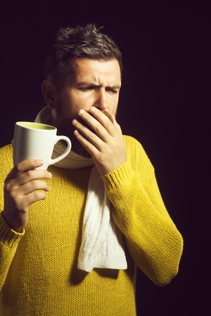 Ill sick bearded man in sweater and scarf holding cup of tea while having cough. Man with flu and fever wrapped in scarf holding cup of healing tea and coughing.の写真素材