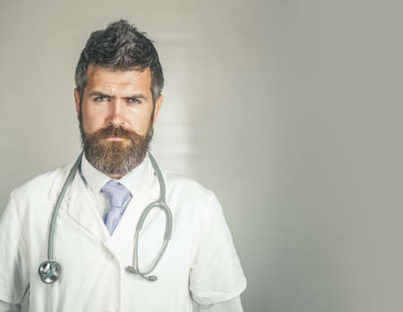 Serious doctor with stethoscope in clinic in surgical uniform. Physician with serious face ready to diagnose. Handsome bearded doctor in white coat. Healthcare and treatment concept.の写真素材