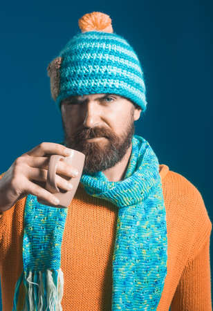 Handsome bearded man in hat, scarf and sweater, holds cup with hot coffee or tea. Attractive caucasian man with beard and moustache in hat and scarf enjoyed drink.の写真素材