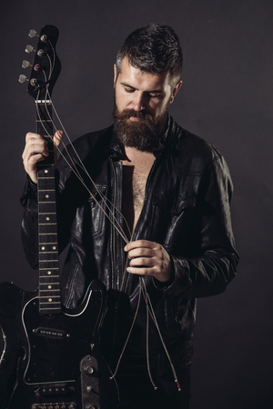 Bearded man, guitarist in black leather jacke with electric guitar. Hobby, music instrument, education, entertainment, concert and learning concept. Charismatic and stylish man holds guitar strings.の写真素材