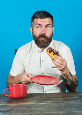 Satisfied bearded man sitting at table with cup of morning coffee. Morning breakfast of coffee and cake at desk. Leisure, food, breakfast concept - handsome man eating tasty pie, having cup cappuccinoの写真素材