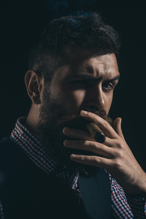 Bearded macho with cigar. Gentleman in checkered shirt and waistcoat smoking cigar. Elegant man with beard, mustache smoke cigar. Classical vintage male portrait. Bearded man in stylish retro clothes.の写真素材