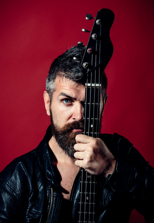 Man with serious face in black clothes holds guitar. Musician, artist play, electric guitar, musical instrument. Man with beard and mustache covers half of face with guitar neck. Hobby, music concept.の写真素材