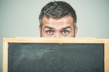 Bearded man hiding behind a black board. Cropped photo of man's head with eyes looking at camera. Man holds black board, ready for your text or advertising. Man holding a chalk board.の写真素材