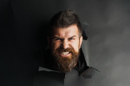 Angry male head looking through paper hole. Portrait angry man peeping through black paper hole. Handsome bearded man watches head out of hole in wall.Copy space for advertising, to insert text,sloganの写真素材