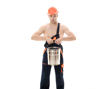 Painter in overalls hold paint bucket. Confident painter, decorator with color bucket. Repair concept. Sexy muscular laborer in protective helmet in overalls. Builder, industrial worker in coveralls.の写真素材