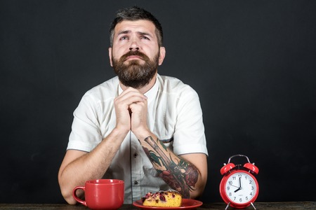 Morning breakfast. Handsome man in white shirt sitting at desk eating breakfast. Grieved bearded man having cup cappuccino, coffee or tea and piece of tasty pie on plate, folded his hands and praying.の写真素材