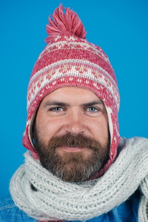 Men winter fashion. Handsome trendy man wearing knit hat and scarf. Portrait of smiling bearded man with scarf and hat on blue background. Young man with scarf and hat in autumn, winter time.の写真素材