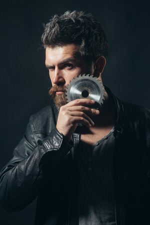 Bearded man, hipster with beard and moustache in black leather jacket and gray sweater holds saw-blade near beard, isolated on dark background. Man with circular saw. Closeup portrait.の写真素材