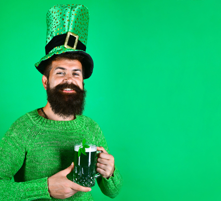 Saint Patrick's Day. Happy bearded man in green sweater holds glass of beer. Clover in glass of beer. Portrait of happy man. Bearded leprechaun. Happy Irish leprechaun with black beard.の写真素材