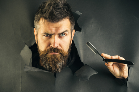 Barbershop concept. Bearded man look through paper. Serious bearded man holds razor. Razor in hands. Barber shop for mens.の写真素材