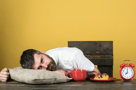Exhausted man resting at table on pillow with alarm clock and cup of coffee on kitchen. Tired bearded man sleep at table after long hours at work. Overworked businessman sleep on desk at breakfast.の写真素材