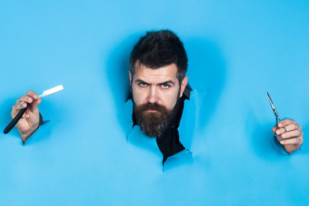 Barbershop concept. Bearded man look through paper. Serious bearded man holds razor and scissors, looking through paper. Razor in hands. Barber shop for mens. Barber shop equipment.の写真素材