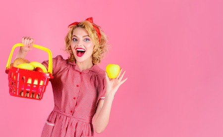 Woman shopping at supermarket. Pinup girl with shopping basket&apple. Smile lady holds grocery basket. Products. Girl puts purchase in basket. Shopping in store/supermarket. Basket household shopping.の写真素材