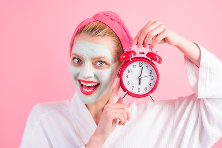 Woman with mask on face holds clock. Beauty Treatments. Facial clay mask. Spa. Cosmetic mask. Beauty mask. Spa treatments. Health. Clean skin. Time. Morning. Morning routine.の写真素材