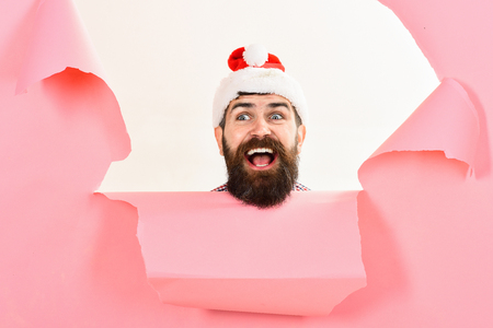 Bearded man in santa hat looking through hole in paper. Christmas, New Year, holidays, winter concept. Stylish guy in santa hat.の写真素材