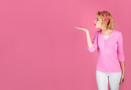 Beauty pin up girl showing empty copy space on open hand palm for text. Concept of advertisement product. Proposing product. Advertisement gesture. Woman in pink sweater raised hand and blowing kiss.の写真素材