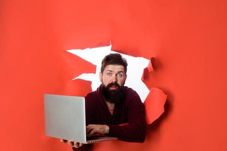 Bearded man through hole in paper holds laptop. Surprised bearded man making hole in paper with laptop. Man with computer. Handsome businessman holds laptop typing, using touchpad. Modern technologiesの写真素材