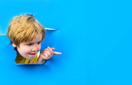 Curious child looks through hole paper. Boy peeking from ripped paper hole. Surprised kid points on copy space. Blonde small toddler shows finger on copy space. Sale, discount, season sales. Advertiseの写真素材