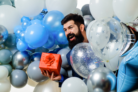 Happy businessman with helium balloons&present box. Stylish bearded man with inflatable air balloons holds gift box. Happy birthday, holidays, celebration, party concept - man with red present box.の写真素材