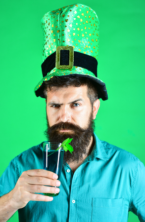 St Patricks green shamrock floating in beer. Green hat with clover. Bearded leprechaun holds green beer. Bearded man in blue t-shirt and green men hat or top hat.の写真素材