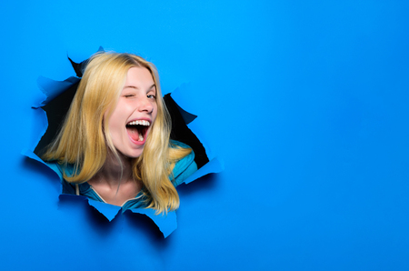 View of woman face through hole in blue paper. Winking smiling girl making hole in paper. Fashion and beauty concept. Copy space for advertising, to insert text or slogan. Discount, sale, season salesの写真素材