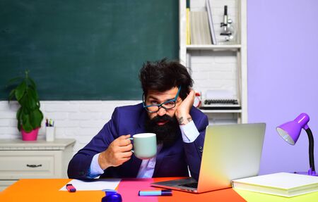 Bored teacher preparing for university exam. Educational process. Tired teacher works with laptop. Student learning in auditorium. Male student thinking about coursework. Sleepy student drinks coffee.の写真素材