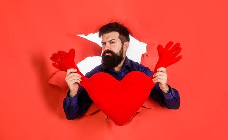 Bearded man holds red heart. Love, Valentines day, holidays, celebrating concept. Romantic man holds heart shape. Businessman holds love symbol. Man with red heart shaped pillow looking through hole.の写真素材