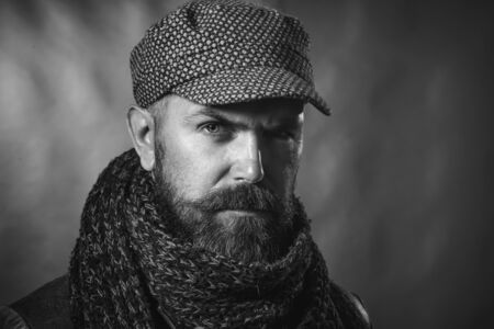 Perfect style. Brutal caucasian hipster wearing warm scarf, cap and jacket. Serious man with beard. Advertising autumn-winter fashion. Attractive macho with beard and mustache. Black and whiteの写真素材
