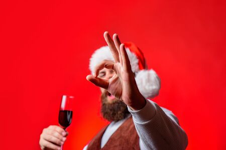 Cheerful santa man holds red wine glass shows ok sign. Santa Claus man doing gesture ok. Bearded man gets ready to celebrate. Winter holiday. Man in Santa hat shows ok gesture. Selective focus on handの写真素材