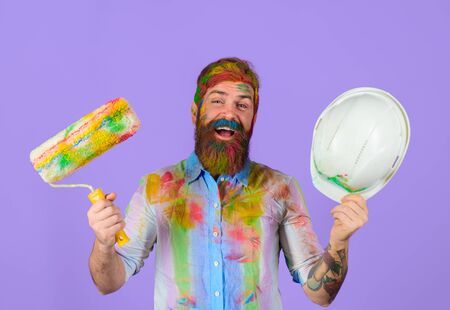 Repair. Painter. Happy bearded painter holds paint roller and helmet. Painter with painting roller. Room painting. Man worker. Construction. Industrial concept. Repair tool. Hardhat. Hard hat in hand.の写真素材