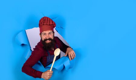 Bearded cook with wooden spoon looking through paper. Chief man in cook uniform holds wooden kitchen cooking tools. Bearded man chef preparing to cook food. Handsome chef cook with kitchen utensilsの写真素材