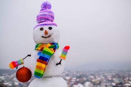 Snowman holds new year ball. Snow man in winter hat. Christmas background with snowman. Funny snowman in stylish hat and scarf. Smiling snow man with Christmas toy. Merry Christmas and Happy new yearの写真素材