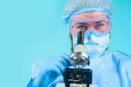 Doctor in protective mask and gloves examining sample through microscope. Male medical doctor using microscope in laboratory. Scientist with microscope making test or research in clinical laboratoryの写真素材