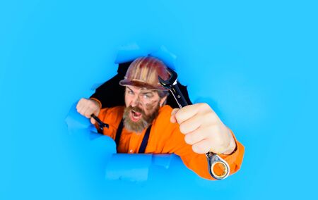Worker in protective helmet breaking paper. Builder holds wrenches. Repairment tools. Spanner. Bearded builder man with wrenches looking through paper. Man builder with wrenches looking through paperの写真素材