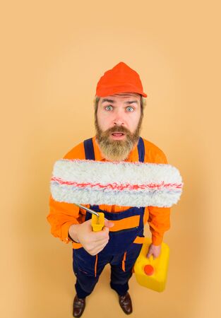 Surprised repairman in uniform holds paint roller. Repair man full height. Professional painter. Designer, builder, worker in hard hat. Painting concept. Bearded man in working clothes with paint rollerの写真素材