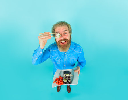 Sushi. Bearded man with sushi near face. Japan. Hipster eating sushi. Sushi delivery. Japanese food. Pickled ginger. Plate of maki rolls. Susi. Exotic nutrition. Sea food. Full height. Copy spaceの写真素材