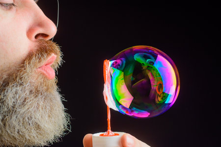 Soap bubbles. Play with bubbles. Bearded man blowing soap bubbles. Happiness. Good mood. Childhood. Closeup portrait of man blowing bubblesの写真素材