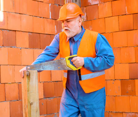 Carpenter at construction site cutting wood with saw. Professional worker in hard hat and overalls.の写真素材