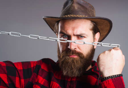 Brutal man in cowboy hat with chain. Serious bearded male in plaid shirt trying to break metal...