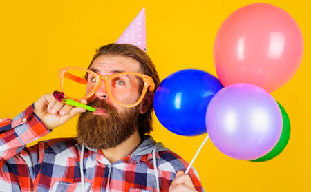 Bearded man in party hat and glasses with balloons blowing noisemaker. birthday celebrationの写真素材