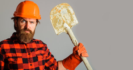 Serious builder in hard hat with shovel. Bearded workman in work gloves and helmet with spadeの写真素材