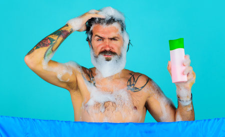 Bearded man in shower with shampoo bottle. Handsome guy with foam on head washing hair. haircareの写真素材