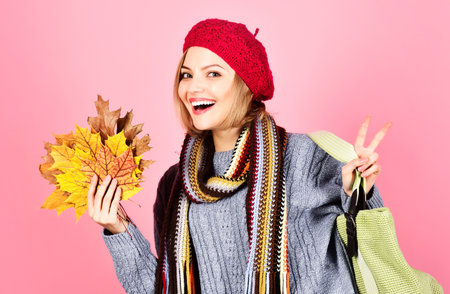 Smiling woman with shopping bag and yellow leaves in sweater, hat and scarf shows sign peaceの写真素材